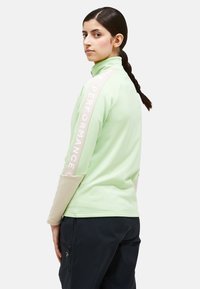 Giacca da performance verde chiaro con colletto alto e maniche lunghe; dettagli beige e testo lungo una manica. Materiale morbido e elastico.