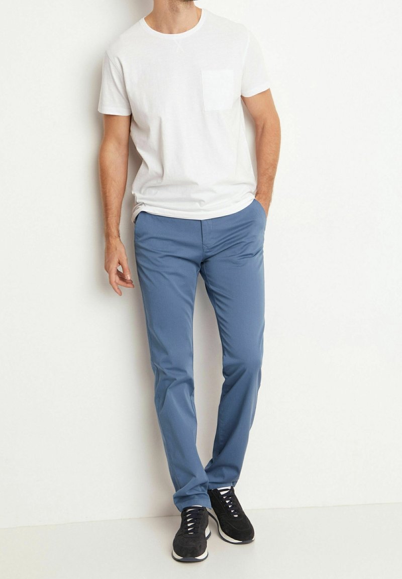 Bílý tričko s kulatým výstřihem a kapsou na hrudi, spárované s modrými elastickými kalhotami a černými teniskami s bílými detaily. Jednoduchý a funkční design.