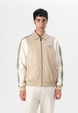 Beige leather bomber jacket with white and olive green sleeves, ribbed cuffs, and a logo patch on the chest. Zipper front closure.