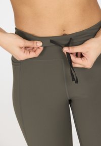 Donkergroene sportbroek met een hoge tailleband, voorzien van een trekkoorddetail. Glad textuur met platgestikte naden langs de zijden.