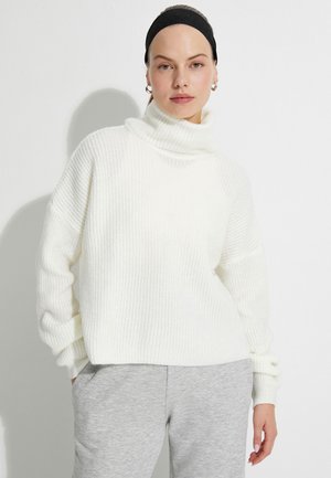 Vit stickad polotröja med ribbat mönster, avslappnad passform och överdimensionerade ärmar, kombinerad med gråa sweatpants.