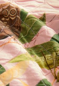 Bunt gemusterte Decke mit einem Schmetterlings- und Blattdesign auf einem rosa Hintergrund, mit sichtbaren Stichen und einer weichen Stofftextur.