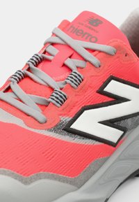 Chaussure de sport rose vif et grise, avec une surface texturée, des lacets gris, et un logo "N" noir et blanc bien en évidence.