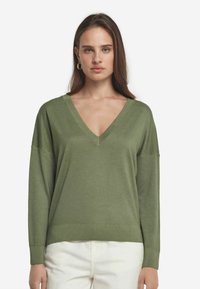 Grön långärmad V-ringad tröja i mjukt material, med avslappnad passform och ribbade mudd, tillsammans med vita byxor.