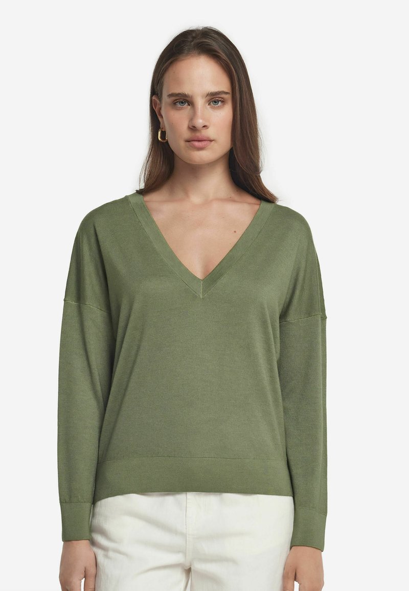 Grön långärmad V-ringad tröja i mjukt material, med avslappnad passform och ribbade mudd, tillsammans med vita byxor.