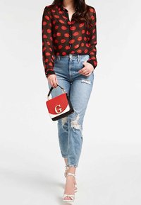 Blouse noire avec un imprimé de lèvres rouges, jeans bleus déchirés taille haute, et un petit sac à main rouge et noir. Le mannequin porte des sandales blanches à lanières.