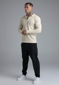 Muškarac s tetovažama u krem majici dugih rukava, crnim hlačama i bijelim tenisicama stoji sa sklopljenim rukama na jednostavnoj sivnoj pozadini.