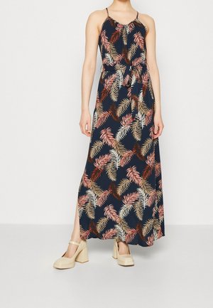 Femme portant une robe maxi sans manches bleu marine avec des imprimés de feuilles roses, beiges et marron et des chaussures Mary Jane beiges à talons blocs.