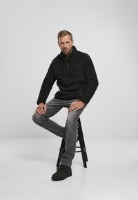 Pullover di pile nero con colletto alto, tasca con zip e superficie testurizzata, abbinato a jeans grigi e stivali neri stringati, seduto su uno sgabello.