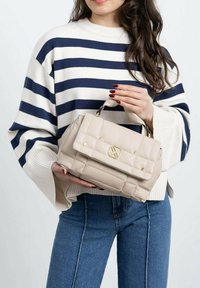 Bolso acolchado beige con forma estructurada, acentos dorados y un asa superior, sostenido por una persona con un suéter a rayas y jeans.
