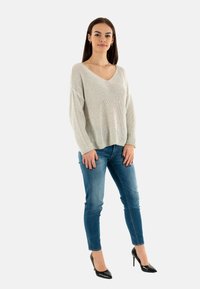 Suéter de punto gris claro con escote en V, corte holgado y mangas largas, combinado con jeans ajustados azules y tacones altos negros de punta.