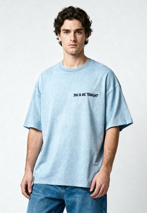 Jeune homme aux cheveux bouclés foncés portant un t-shirt oversize bleu clair avec le texte "YOU & ME TONIGHT" et un jean bleu sur un fond uni.