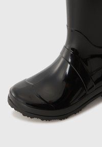 Botte de pluie noire brillante avec semelle texturée et détails de couture subtils sur un fond clair.