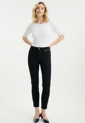 Jeans ajustados negros con un detalle de cremallera dorada en los bolsillos frontales, combinados con una blusa blanca de canalé ceñida y tacones negros de punta.