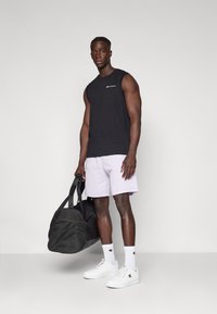 T-shirt sans manches noir, shorts violets clairs, baskets blanches, chaussettes blanches avec logo. Tenant un sac de sport noir avec un panneau en maille.