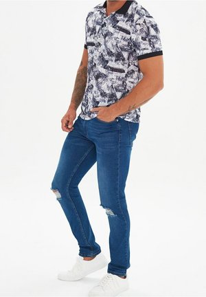 Mand iført hvide sneakers, slidte blå skinny jeans og en sort-hvid poloshirt med abstrakt mønster, stående med den ene hånd i lommen.