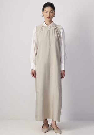 Abito beige senza maniche con cerniera, caratterizzato da un scollo raccolto e una silhouette dritta. Indossato sopra una blusa bianca. Scarpe flat.