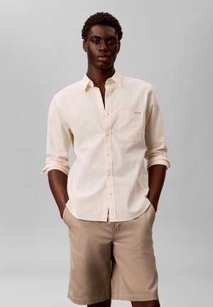 Jeune homme portant une chemise crème à manches longues boutonnée et un short beige, debout les mains dans les poches sur un fond gris uni.