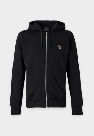Jeune homme debout portant un sweat à capuche noir zippé, un t-shirt blanc, un jean bleu et des chaussures marron sur un fond clair uni.