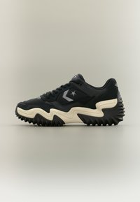 Μαύρο αθλητικό παπούτσι με μπεζ κυματιστή σόλα, λογότυπο αστέρι και σήμα chevron στο πλάι, υφασμένο πέλμα και επάνω μέρος από δίχτυ και σουέτ σε απλό φόντο.