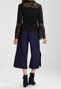 Blouse noire avec détails en dentelle sur les manches et le dos, associée à un pantalon large bleu marine avec fentes latérales. Bottines noires complètent le look.