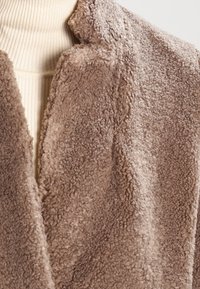 Beżowy teddy polarowy płaszcz częściowo zakrywający kremowy sweter z rdzeniem w prążki na neutralnym tle.