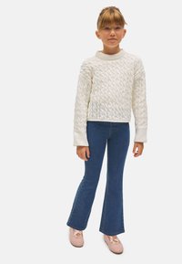 Jersey de punto blanco con cuello redondo, combinado con jeans acampanados azules y zapatos rosa claro. Diseño simple y texturizado.