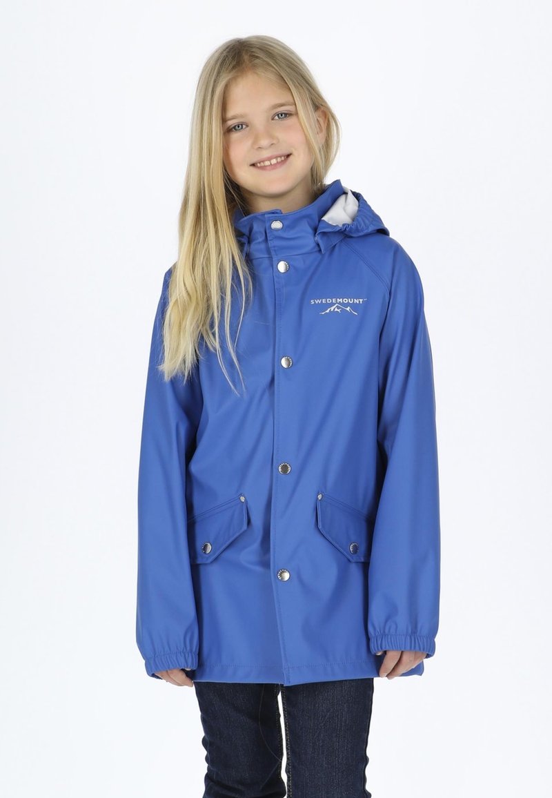 Fille souriante aux longs cheveux blonds portant une veste de pluie bleue Swedemount avec des poches avant et un jean foncé, sur fond blanc.