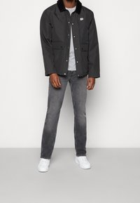 Sort vandtæt jakke med et corduroy-krave, grå T-shirt nedenunder, grå jeans og hvide sneakers, stående foran en lysegrå baggrund.