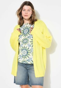 Cárdigan de punto amarillo brillante con mangas largas, abertura frontal y puños acanalados, combinado con una blusa de estampado floral y pantalones cortos de mezclilla.