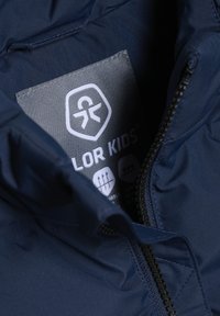 Marineblå isolert jakke med svart glidelås. Den har et stoffmerke som viser vaske-symboler og "COLOR KIDS"-logoen inne i kragen.