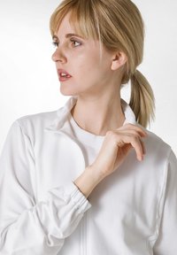 Veste blanche zippée avec un col montant et des poignets élastiques, superposée à un t-shirt blanc à col rond ; texture de tissu lisse.