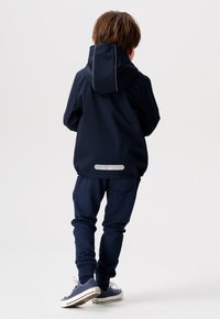 Veste à capuche bleu marine avec des accents réfléchissants, assortie à un pantalon de jogging ; le tout en tissu doux et texturé. Des baskets blanches complètent le look.