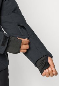 Sort polstret jakkeærme med en åbning og tommelhuller. Har en glat tekstur og velcro-lukning. Hånden justerer ærmekanten.