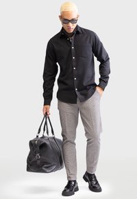 Camisa negra de botones con un bolsillo en el pecho, pantalones grises de cuadros y un bolso de cuero negro. El modelo lleva gafas de sol y zapatos negros, con un pañuelo.