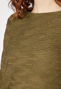 Pulover texturat de culoare verde măslin, cu un model în valuri, având o croială lejeră și un decolteu larg. Țesătura pare moale și ușor transparentă.