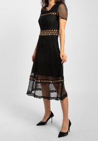 Robe en dentelle noire à manches courtes, présentant un motif géométrique, des accents circulaires dorés à la taille et un ourlet festonné.