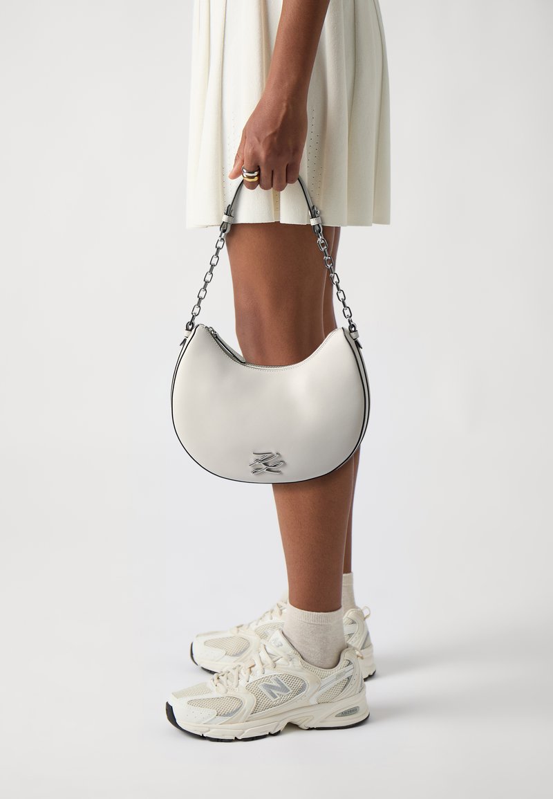 White leather handbag with a crescent shape, zip closure, and silver chain handle. Features a black accent along the edge and logo detail.