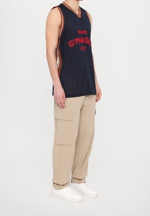 Mann trägt ein marineblaues Basketballtrikot mit rotem Text, beige Cargohosen und weiße Turnschuhe, steht vor einem schlichten weißen Hintergrund.