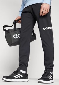 Black sports pants with white "adidas" logo, elastic cuffs, paired with black sneakers featuring white stripes and holding a black gym bag.