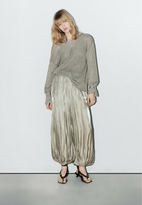 Przesadnie duży beżowy sweter dzianinowy nałożony na błyszczące, plisowane srebrne spodnie z elastycznymi mankietami; modelka w czarnych sandałach na obcasie.