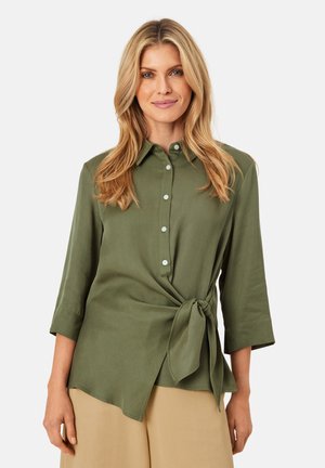 Olijfgroene, knoopsluiting blouse met 3/4 mouwen, gestrikt op de taille. Glad stof, kraag en kleine knopen als detail. Gecombineerd met beige onderkleding.