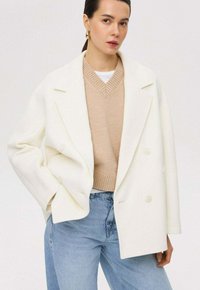 Witte dubbelgebreide jas met een notch-kraag, gedragen over een beige V-hals trui, gecombineerd met lichtblauwe high-waisted jeans.