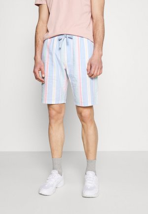 Homme portant un short rayé pastel, un t-shirt rose clair, des chaussettes grises et des baskets blanches, debout devant un fond uni.