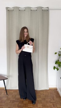 Svarta vida byxor i kombination med en figursydd svart topp. Håller en vit handväska med ett guldspänne. Texturerad beige gardin i bakgrunden.