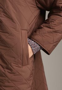 Smešani smeđi kaput s prenaglašenim dijagonalnim uzorkom, s bočnom džepom i širokim rukavima. Unutarnji kraj rukava pokazuje pleteni dizajn u više boja.