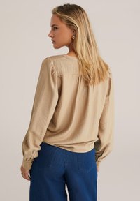 Beige Bluse mit langen Ärmeln und strukturierter Oberfläche, lockerer Passform und gerafften Ärmeln. Kombiniert mit einer dunkelblaue, weit geschnittenen Hose.