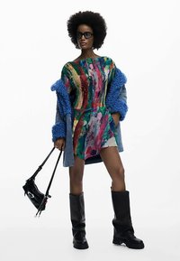 Mehrfarbiges Kleid mit strukturiertem Muster, kombiniert mit einem blauen Kunstpelzmantel. Schwarze hohe Stiefel und eine kleine schwarze Tasche vervollständigen den Look.