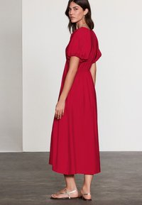 Punainen puuvillainen maksimekko, jossa on puffihihat, vyötärö kavennettu ja liehuva helma. Yhdistetty hopeisiin sandaaleihin, kuvattuna harmaalla teksturoidulla lattialla.