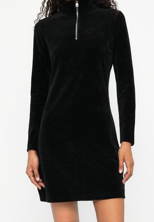 Woman wearing a long-sleeve black velvet mini dress with a front silver zipper and subtle embroidered logo on the chest.
